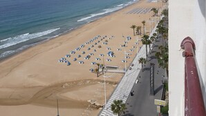 Uma praia com fileiras de guarda-sóis azuis e espreguiçadeiras, um calçadão com palmeiras e um horizonte urbano ao fundo.