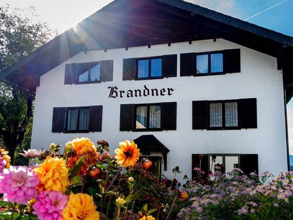 Ein weißes Gebäude mit dem Schriftzug 'Brandner' auf der Fassade, mit zahlreichen Fenstern und einem Garten mit Blumen im Vordergrund.