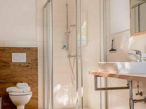 Un baño moderno con ducha de vidrio, un lavamanos blanco sobre encimera de madera y un inodoro con toallas apiladas.
