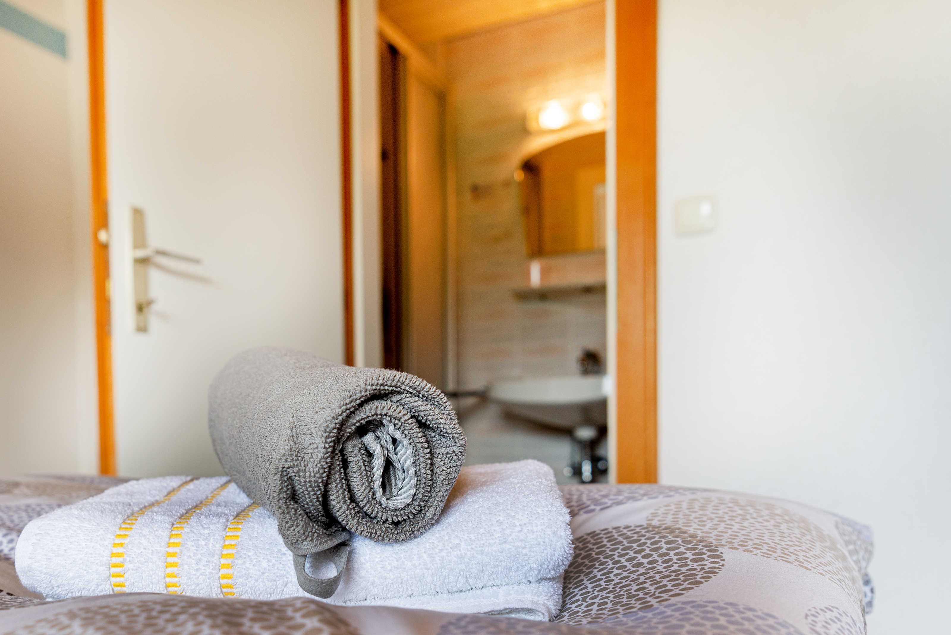 Un lit avec des serviettes enroulées et un coussin à motifs, une porte donnant sur une salle de bain, et un miroir qui reflète la salle de bain.