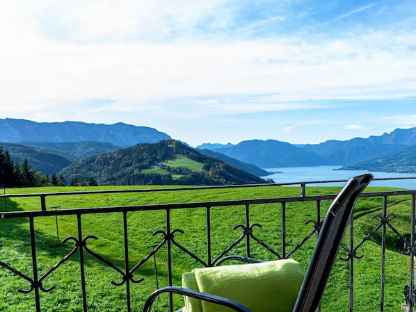 Un balcón con barandilla metálica, una silla verde y vista a un lago rodeado de montañas.