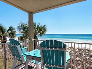 A covered outdoor seating area with two chairs and a table, overlooking a beach with palm trees and the ocean. - Near Water Park, Beach Front, Balcony (Destin)