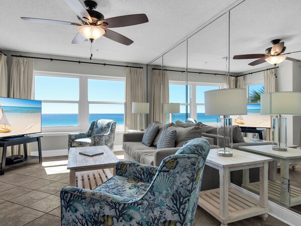 A spacious living room with a large window offering a view of the beach, a ceiling fan, and a modern seating arrangement.