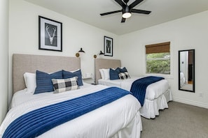 A bedroom with two beds, a ceiling fan, framed artwork on the walls, and a window with a view of greenery.