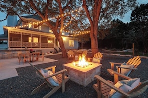 A fire pit surrounded by wooden chairs and a dining table, with string lights and a house in the background. - Hill Country Escape | Pool, Hot Tub & Sleeps 25 (Austin)