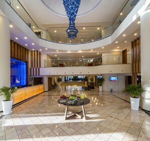 A hotel lobby with a reception desk, a large digital screen, a chandelier, a table with fruit and flowers, and a second-floor balcony. - Kaya Side Hotel (Manavgat)