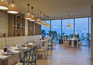 A restaurant with wooden tables, wicker pendant lighting, and large windows overlooking a scenic view. - Kaya Side Hotel (Manavgat)