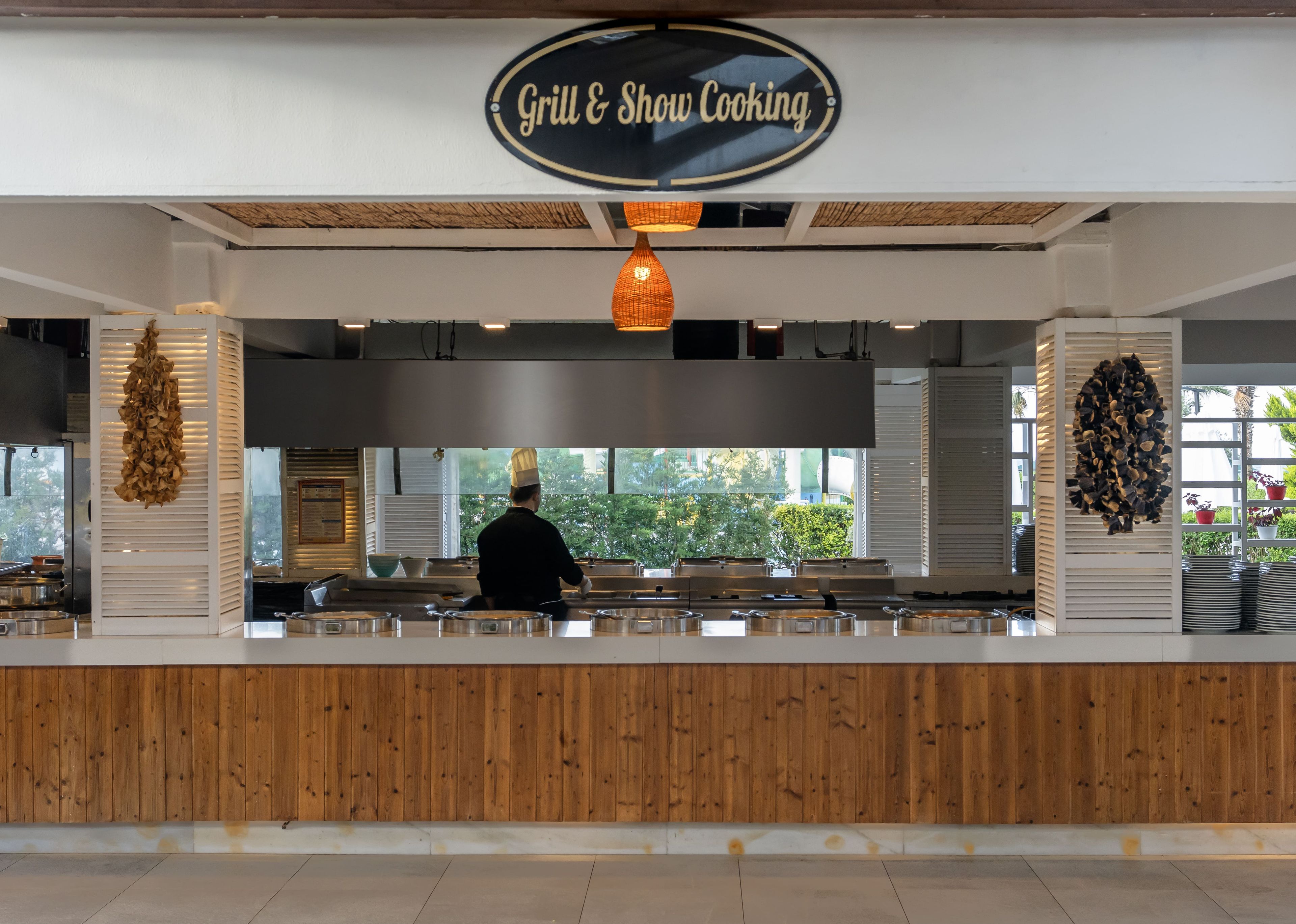 Restaurante com placa indicando 'Grill & Show Cooking', um cozinheiro trabalhando e vista para o verde lá fora.