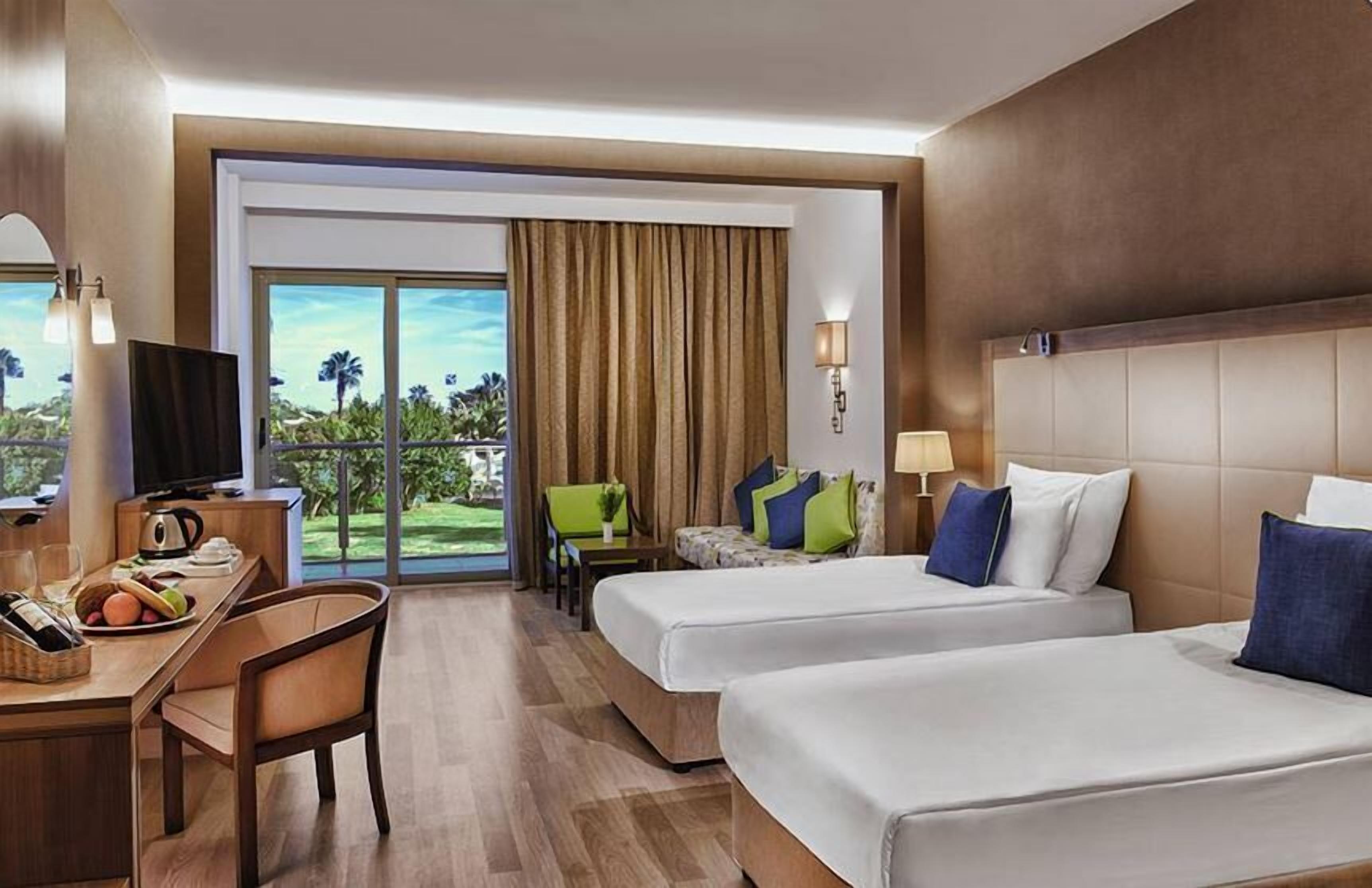 A hotel room with two beds, a desk with a television, a dining area, and a view of the outdoors.