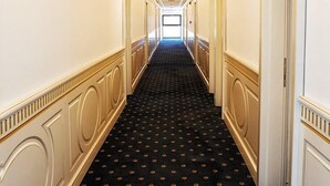 A long, narrow hallway with a black carpet featuring a small, repeating pattern. The walls are white with gold trim and decorative molding. - Hotel Terra (Neptun)