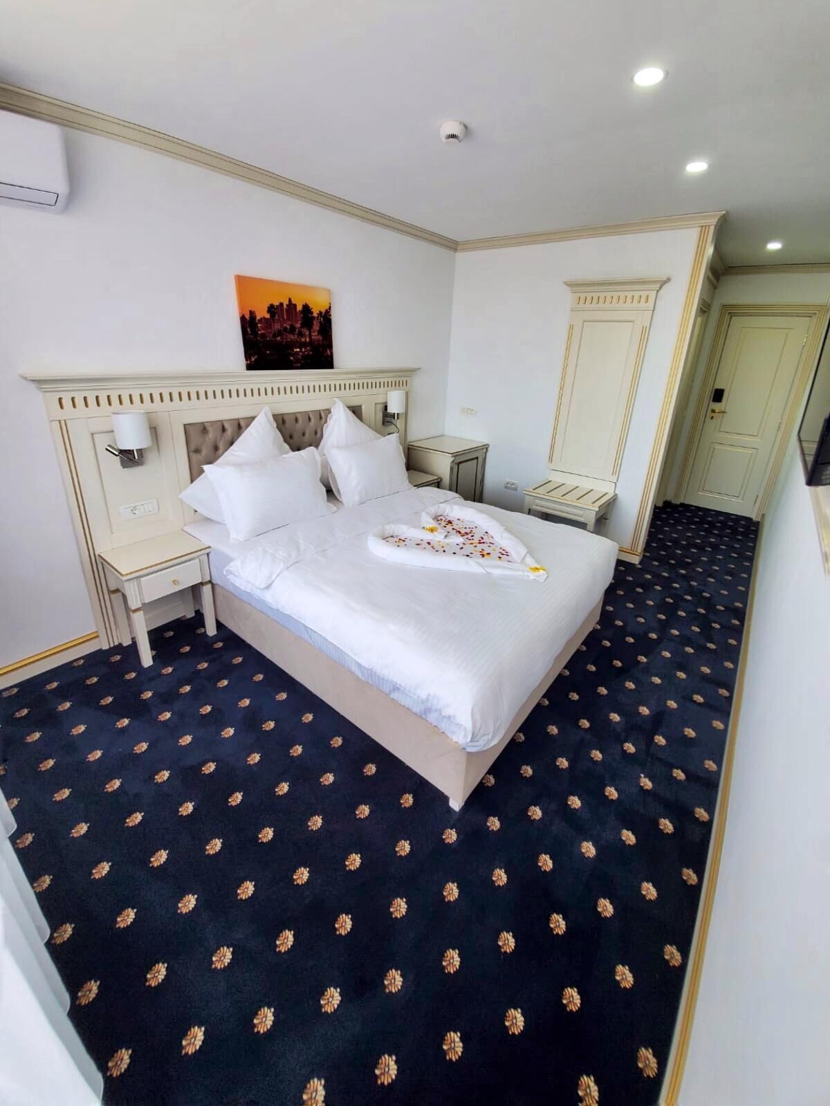 A hotel room with a large bed, a nightstand, a bedside table, a wall-mounted lamp, and a door with a mirror.