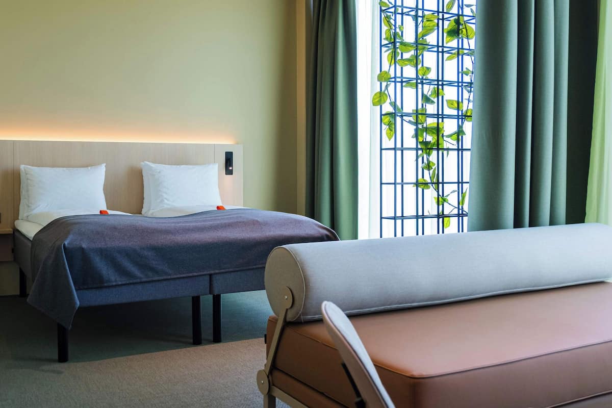 A hotel room with a bed, bedside tables, a bench, and a window with a view of greenery.
