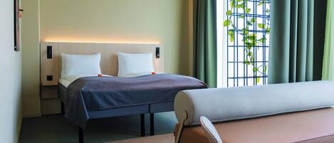 A hotel room with a bed, bedside tables, a bench, and a window with a view of greenery.