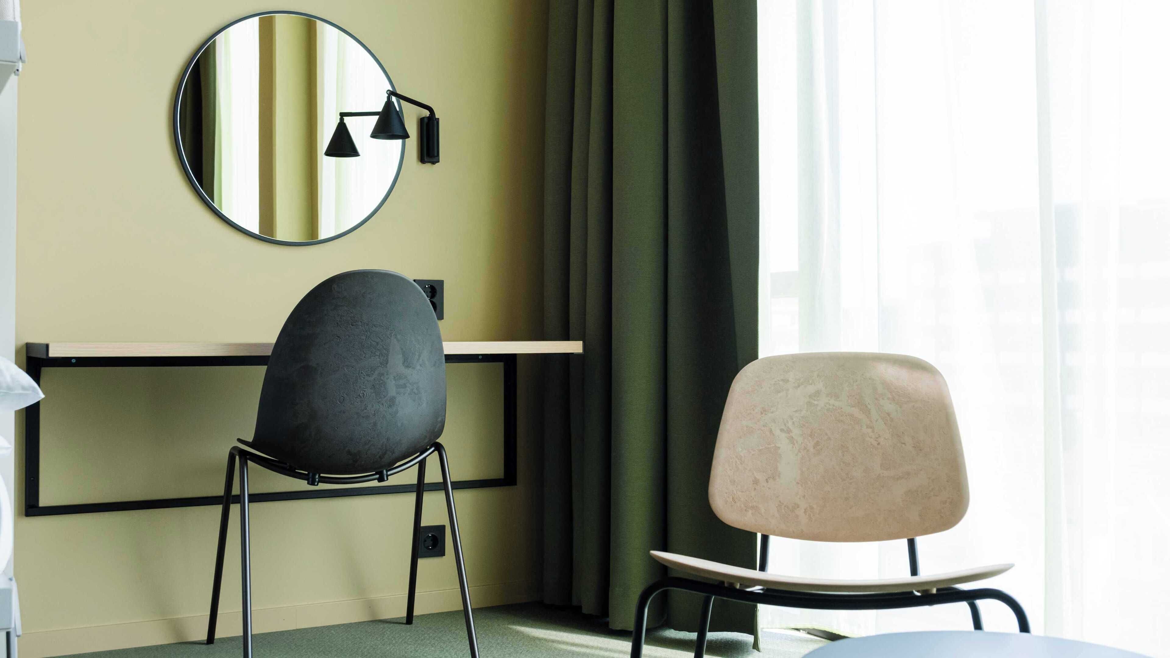 Habitación de hotel con escritorio, dos sillas, un espejo redondo y una ventana con cortinas.