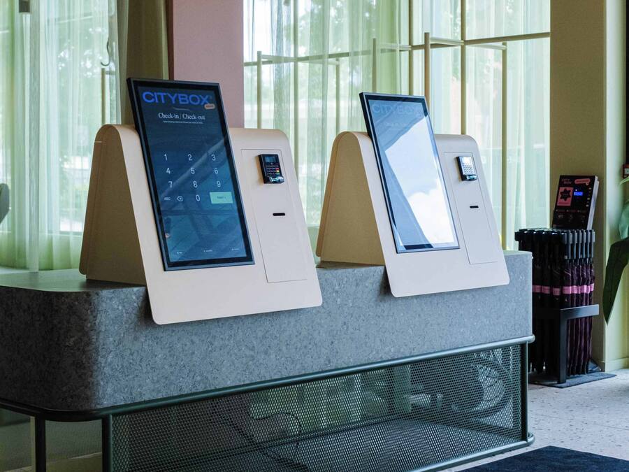 A reception area with two CityBox kiosks for check-in and check-out, a digital sign above the reception desk, and a CityBox mat on the floor.