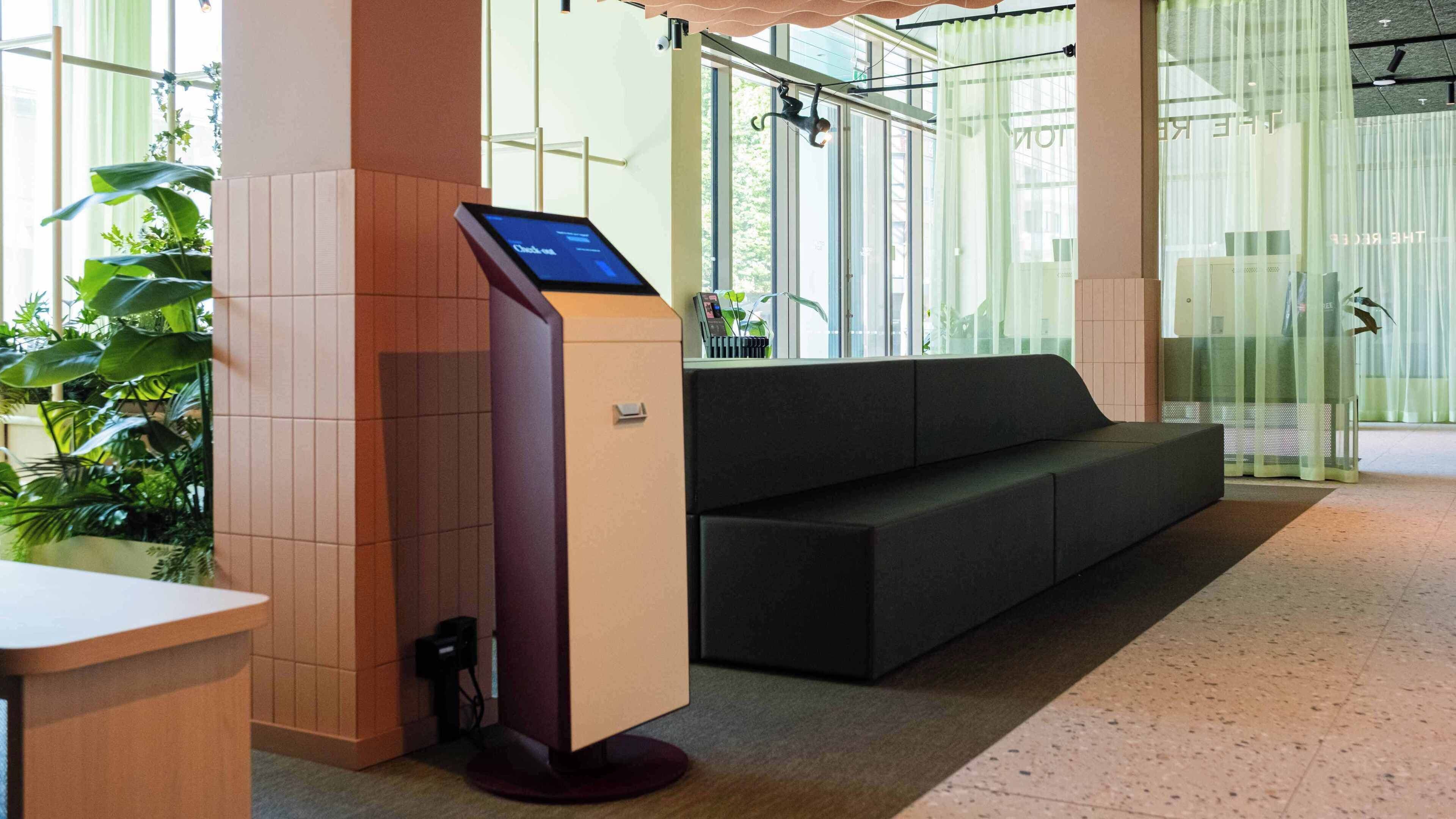 A reception area with a digital kiosk, a sofa, and a plant.