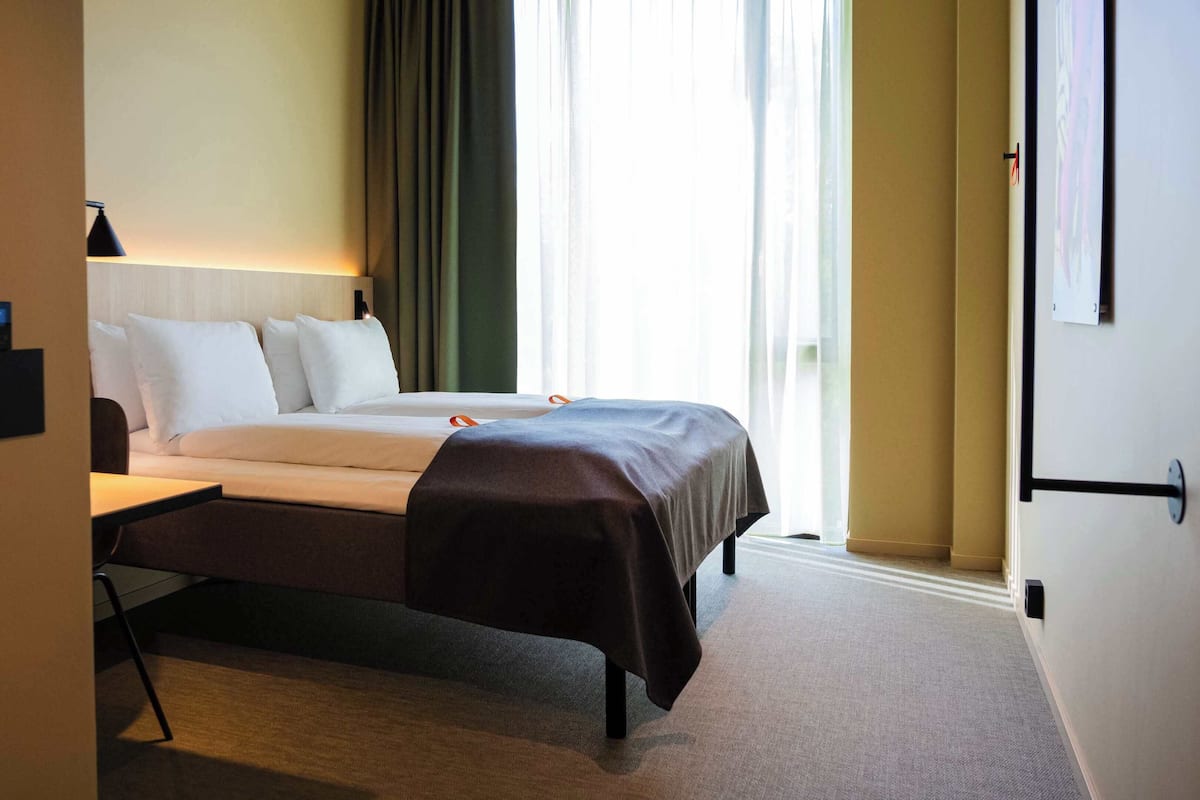 A hotel room with a bed, bedside table, and a window with curtains.