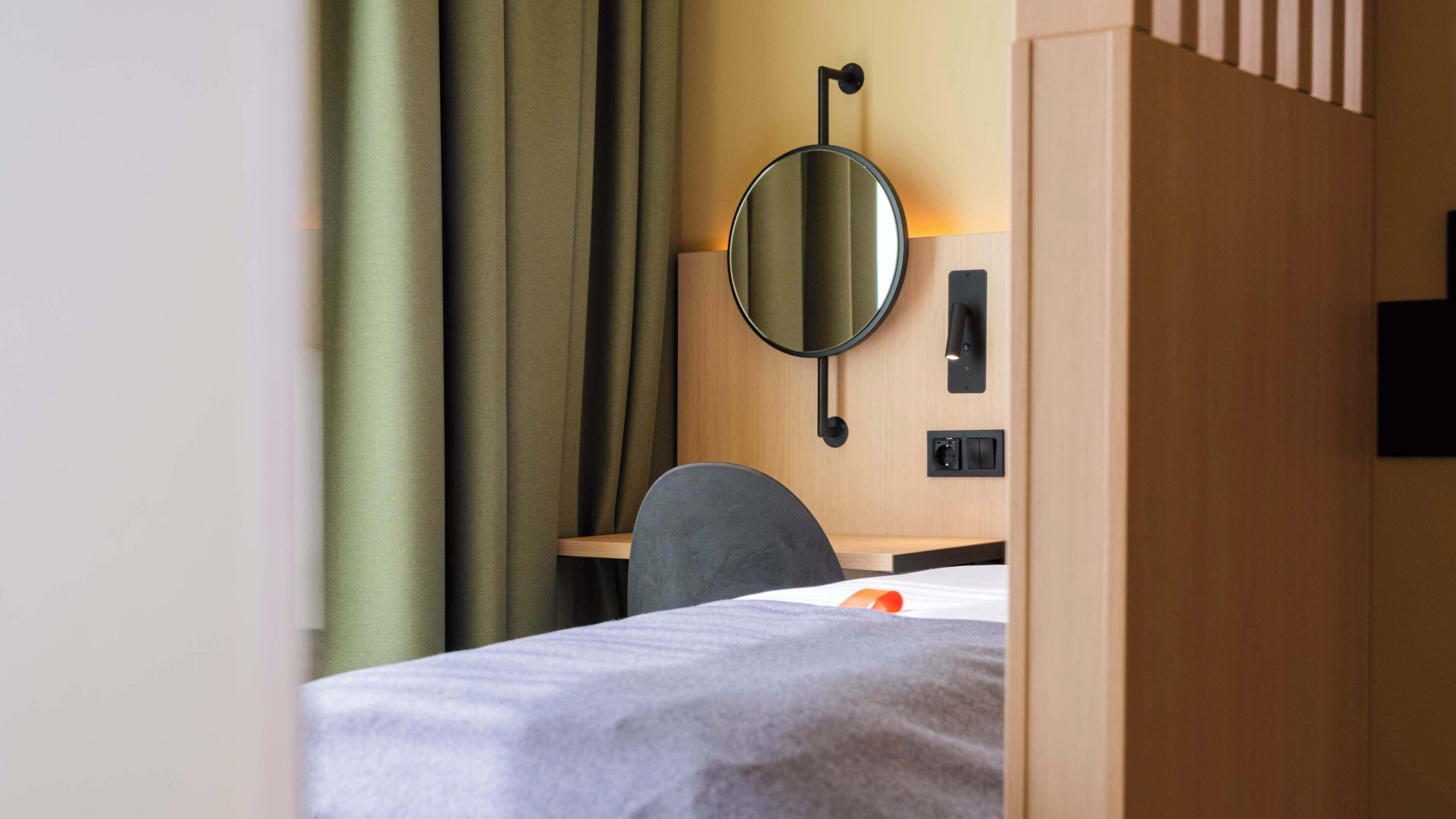 A hotel room with a bed, a desk, a chair, a mirror, and a wall-mounted light fixture.