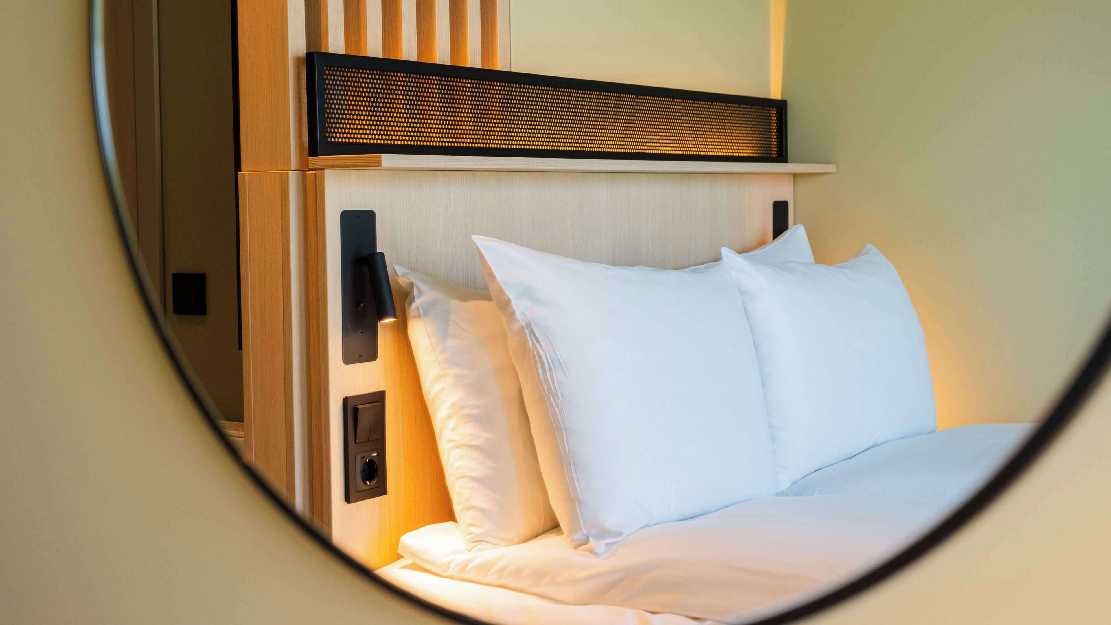 A bedroom with a bed, pillows, a wall-mounted light, and a wooden panel above the bed.
