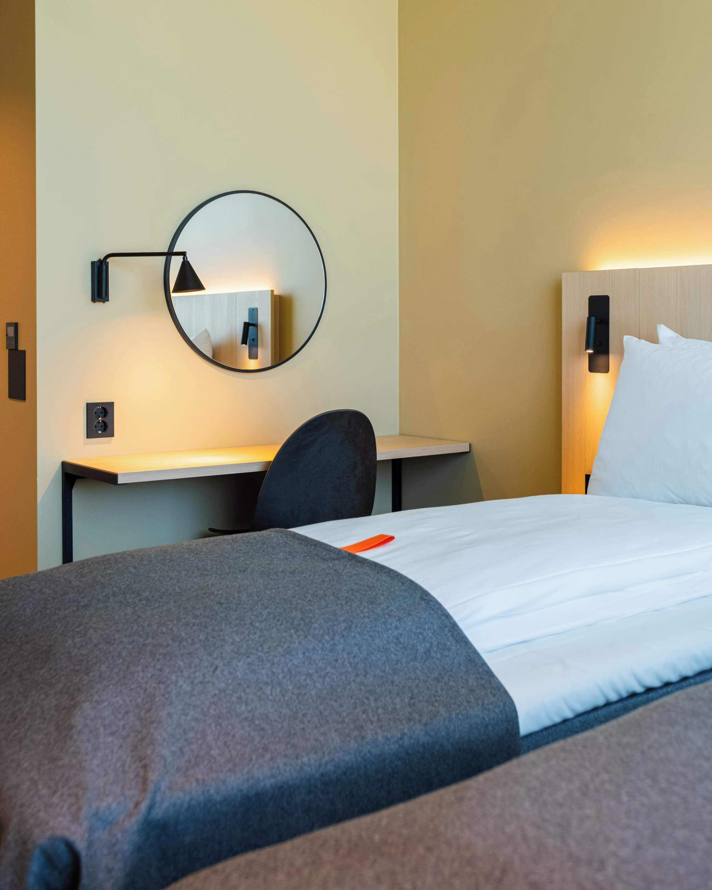 Une chambre d’hôtel équipée d’un lit, d’un bureau, d’une chaise, d’un miroir rond et d’une lampe fixée au mur.