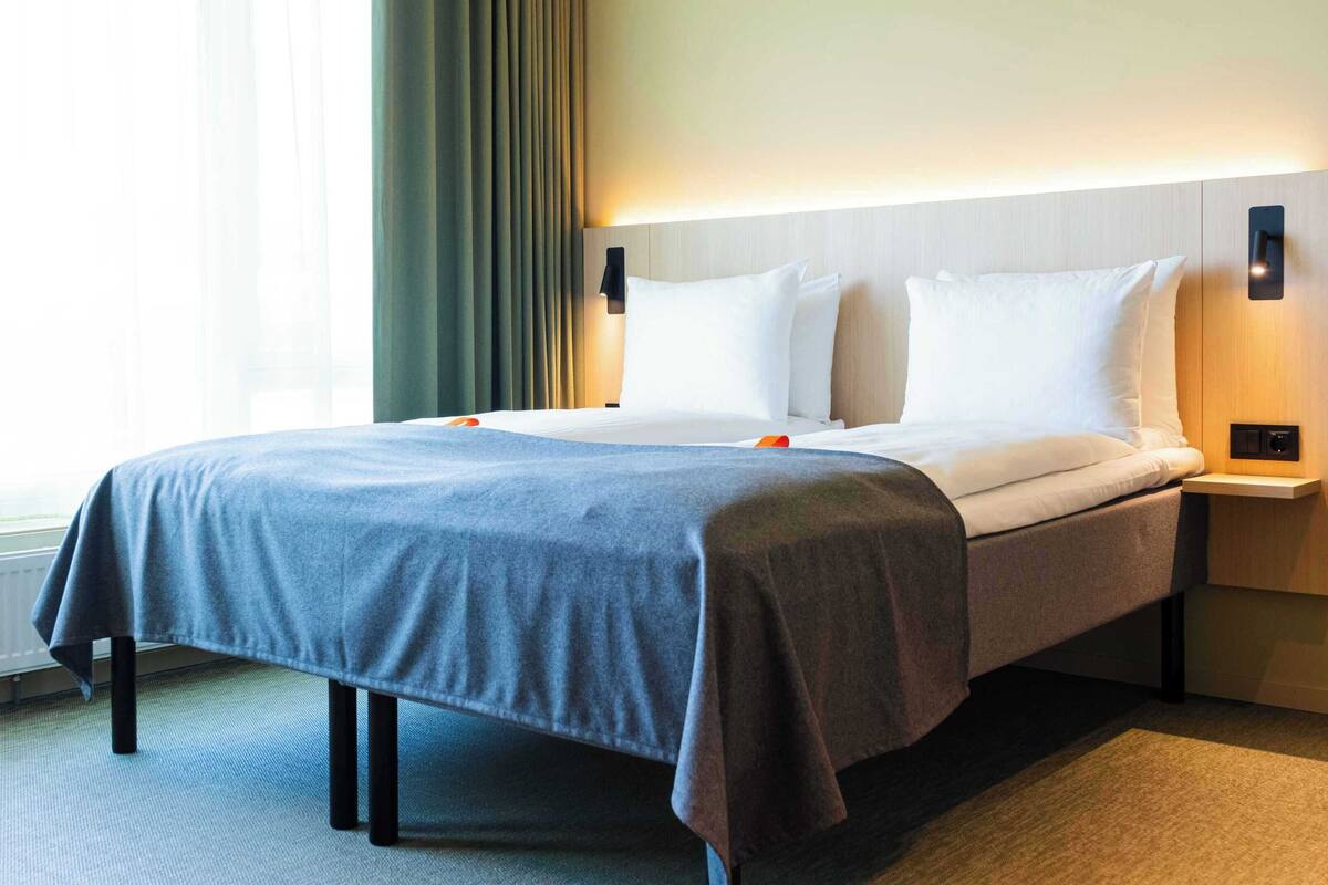 A hotel room with a large bed, a bedside table with a lamp, and a window with curtains.