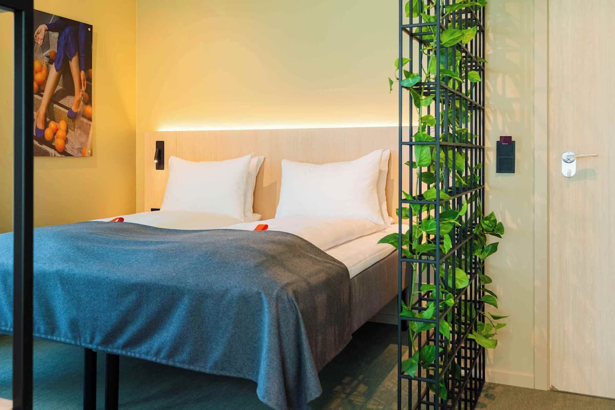 A hotel room with a bed, a blue blanket, white pillows, a wall-mounted light, a painting, and a green plant in a black trellis.