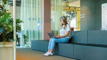 Une personne assise sur un banc gris dans un espace intérieur moderne, devant un ordinateur portable.