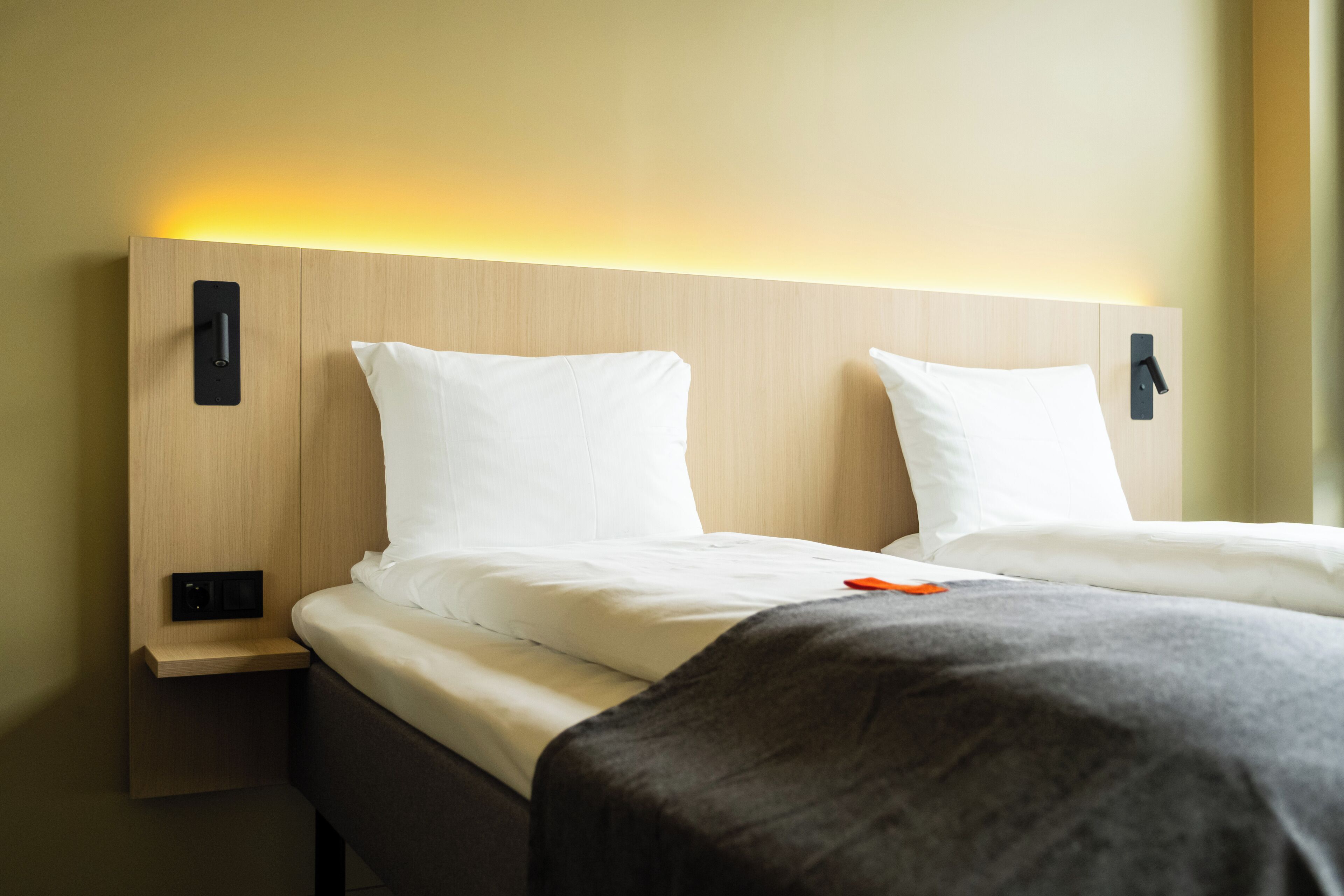 A hotel room with two beds, each with white linens and a headboard. There is a bedside table with a lamp on each side of the beds.