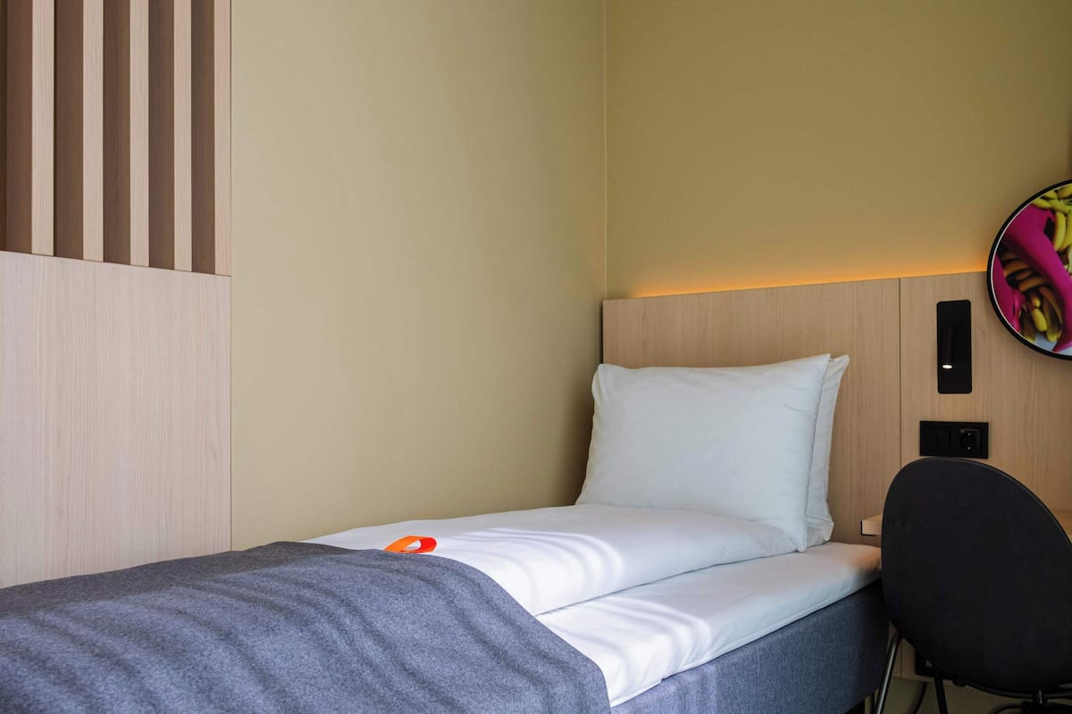 A hotel room with a single bed, a grey blanket, a white pillow, a black chair, and a wall-mounted circular mirror with a banana-themed design.