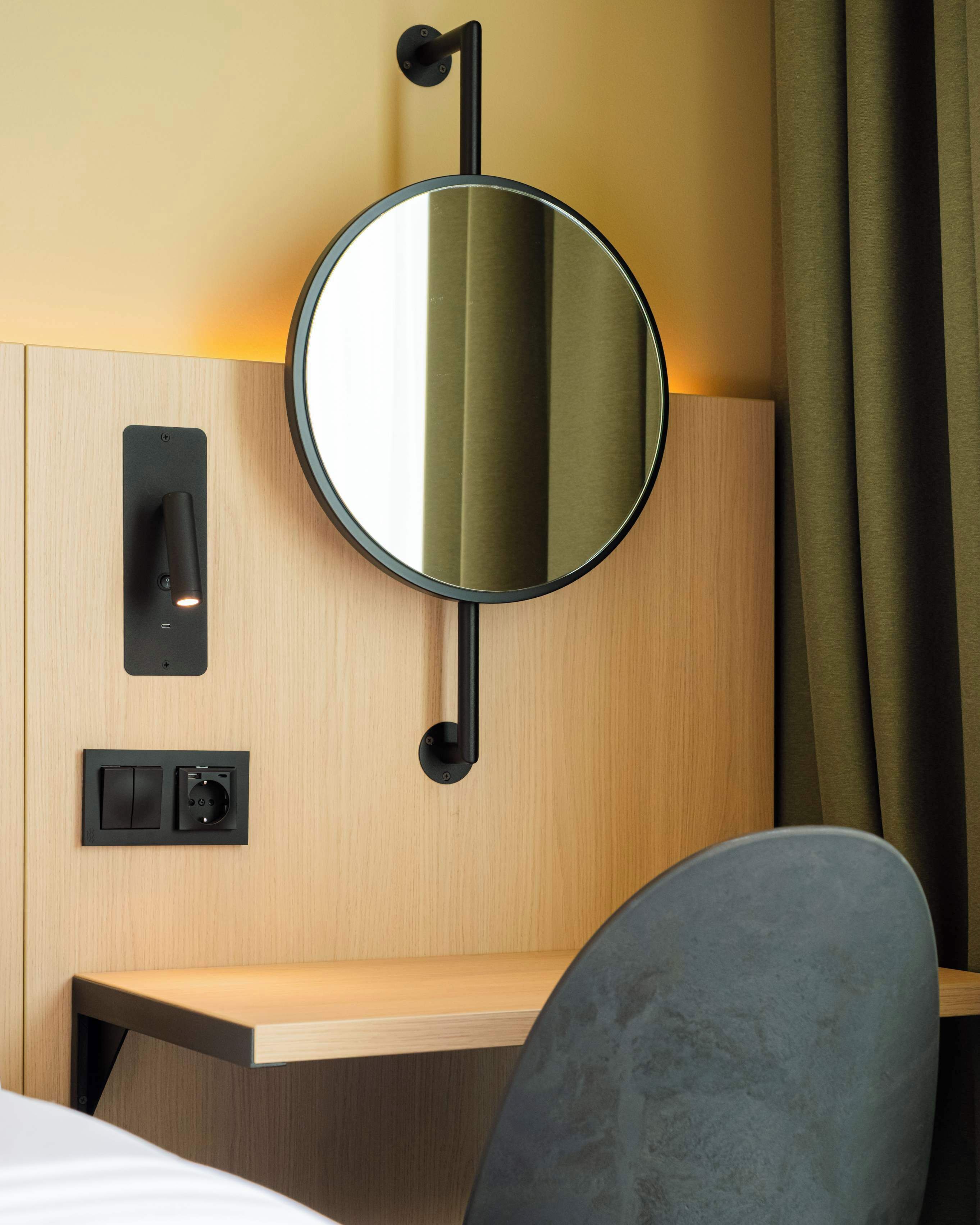 Ein modernes Hotelzimmer mit einem Holz-Kopfteil, einem runden Spiegel, einem Nachttischschalter und einem kleinen Nachttisch.
