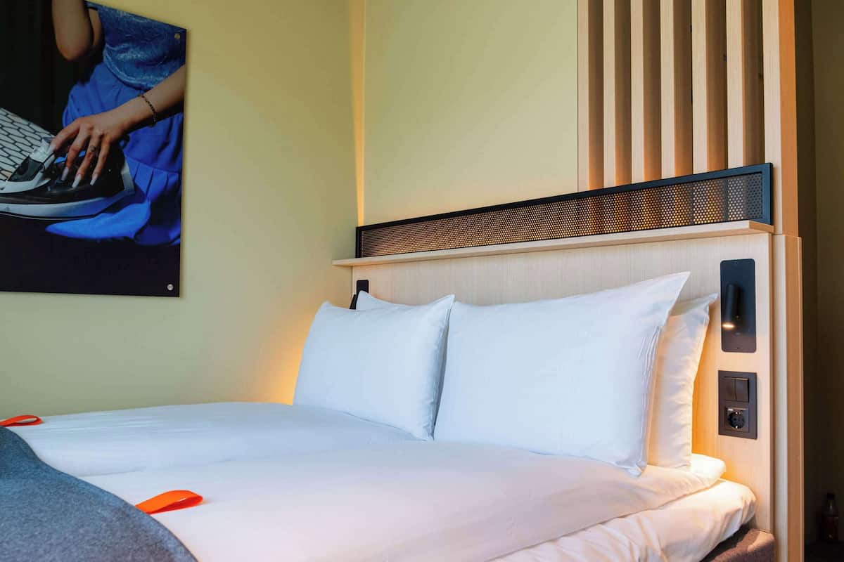 A hotel room with a bed, pillows, a headboard with a wooden panel, a wall-mounted light, and a poster on the wall.