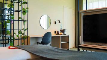 A hotel room with a bed, a desk, a chair, a TV, and a plant.