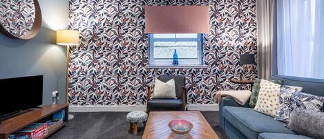 A living room with a patterned wallpaper, a sofa, a coffee table, a TV, and a window with curtains.
