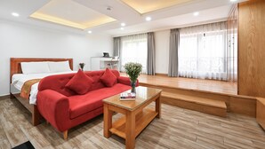 A modern hotel room with a bed, red sofa, wooden coffee table, and a view of the city through large windows.