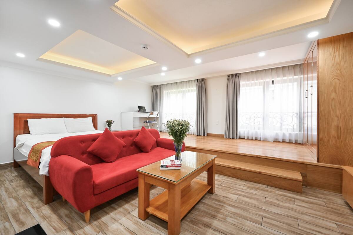 Une chambre d’hôtel moderne, dotée d’un lit, d’un canapé rouge, d’une table basse en bois et offrant une vue sur la ville grâce à de grandes fenêtres.