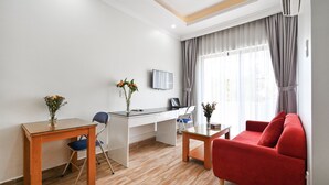 A modern living room with a red sofa, a wooden coffee table, a glass-top dining table with a blue chair, a white desk with a laptop, and a flat-screen TV mounted on the wall.