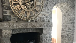 A stone fireplace with a large clock above it, a white leather sofa to the right, and a stone wall with an archway.