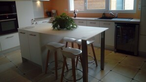 A modern kitchen with white cabinets, a central island with stools, a built-in oven, a coffee maker, and a window with a view of the outside.