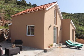 A small building with a tiled roof, a balcony with lounge chairs, and a stone wall in the background.