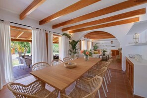 A dining area with a wooden table and wicker chairs, an open kitchen with white cabinets, and a view of the outdoors. - Dream finca: XXL pool, table tennis, BBQ house, volleyball, air-condition (Port de Pollença)