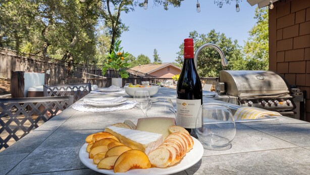 An outdoor dining table with a plate of cheese, bread, and a bottle of wine, a grill, and string lights in the background. - 5 Bedroom wine Country Home, Grill, Hot Tub, Granny unit in Santa Rosa  (Santa Rosa)