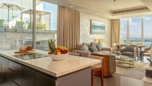 A modern living room with a large window, a dining area, and a kitchen with a fruit bowl.