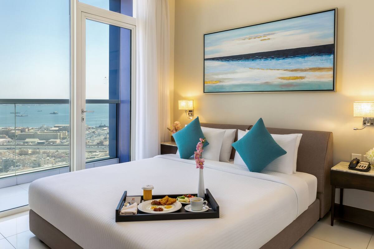 A hotel room with a large bed, a city view, a painting, and a tray with breakfast.