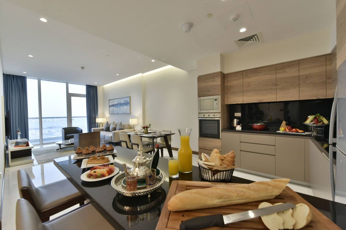 A modern hotel room with a dining area, kitchenette, and a large window offering a city view.