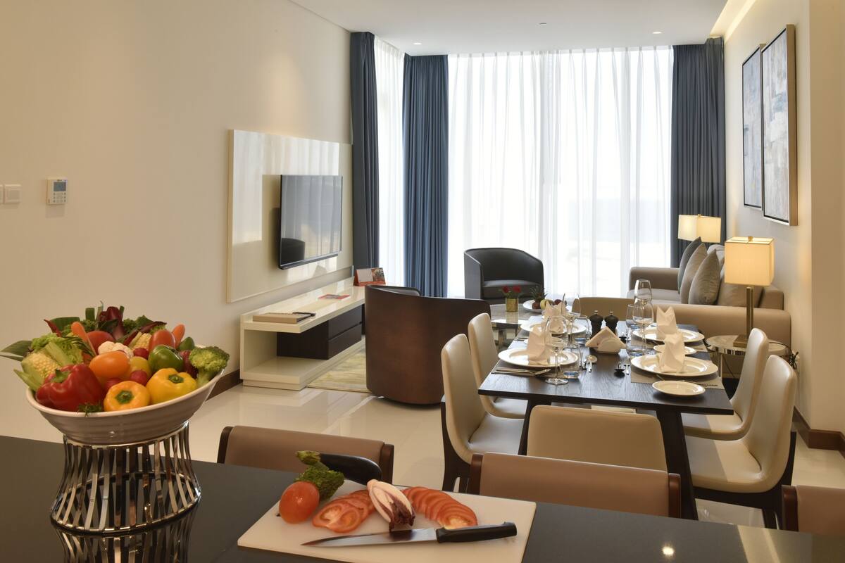 A modern hotel dining area with a table set for two, a bowl of fresh vegetables, and a television mounted on the wall.