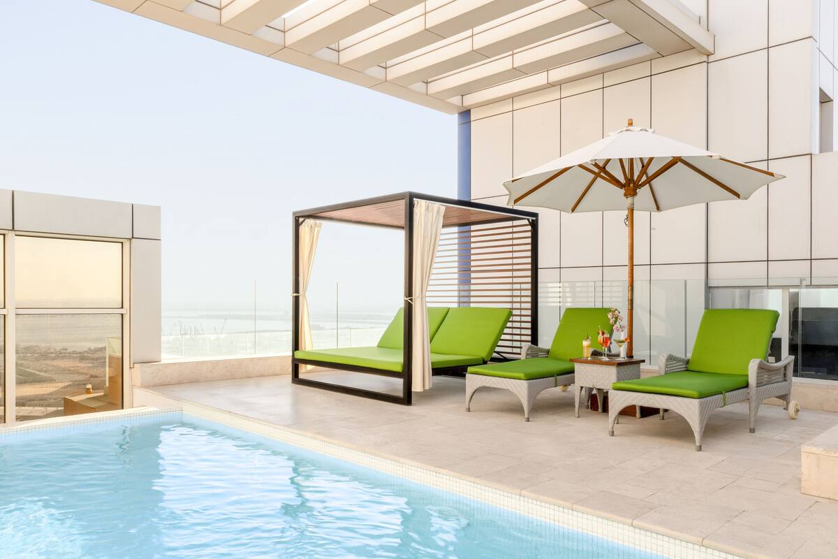 A modern indoor pool area with a glass balustrade, green cushioned lounge chairs, and a large umbrella.
