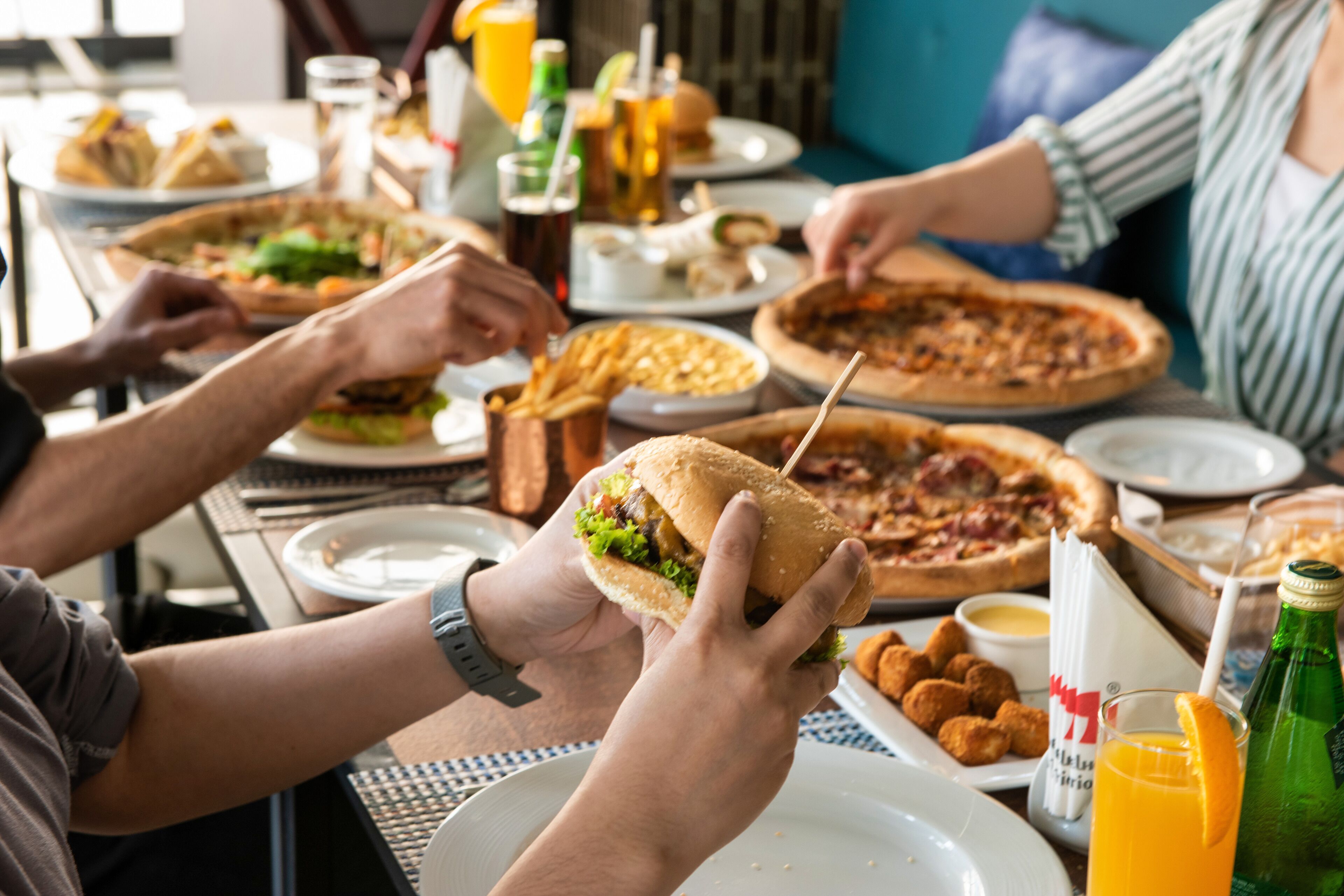 Eine Gruppe von Personen isst in einem Restaurant. Auf dem Tisch befinden sich verschiedene Gerichte, darunter Pizza und Sandwiches.