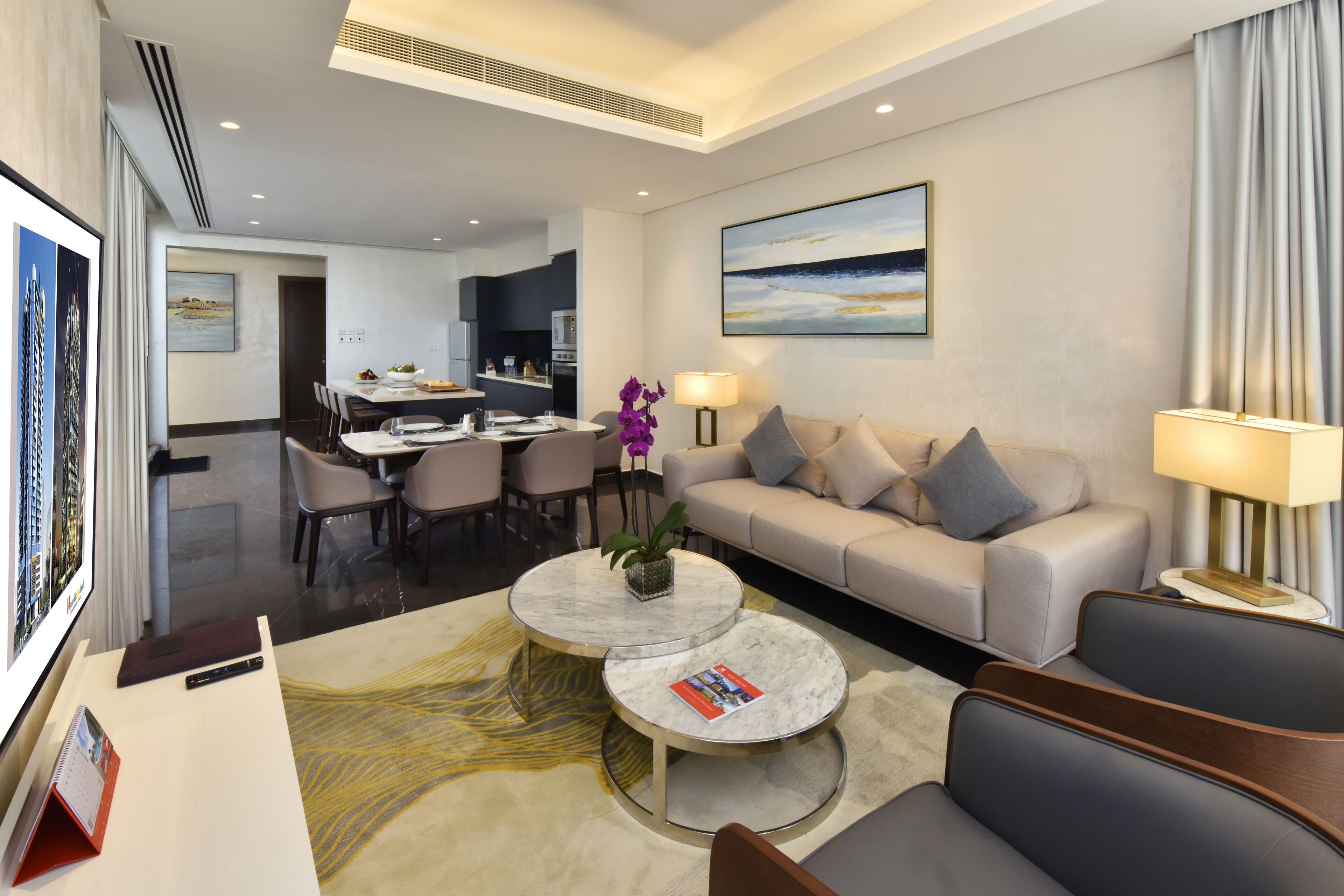 A modern hotel room with a sofa, two round coffee tables, a dining area, and a kitchen.