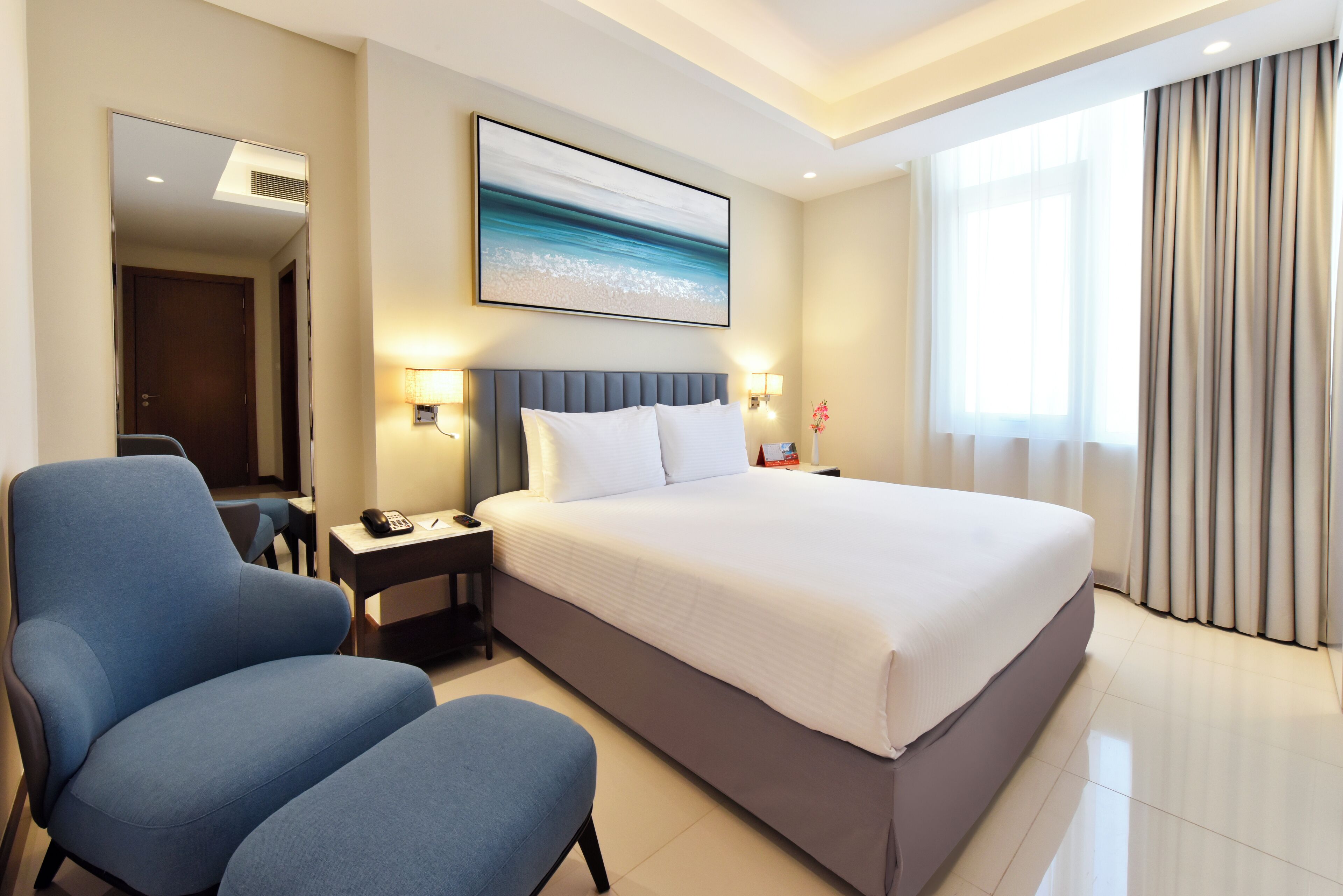 Une chambre d’hôtel avec un grand lit, un fauteuil bleu, une table de chevet avec une lampe et un tableau représentant une scène de plage.