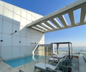 Un espace piscine aménagé sur le toit, avec des chaises longues et une balustrade en verre donnant sur le paysage urbain.